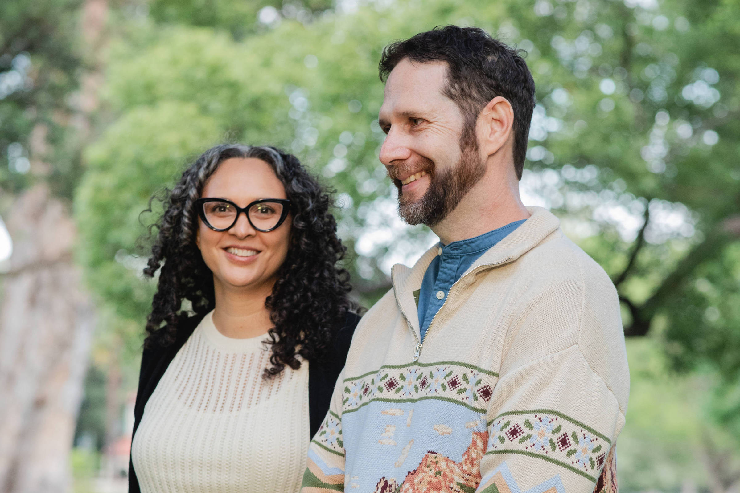 Um casal está posando em um parque. A mulher à esquerda tem cabelo cacheado e usa óculos grandes, vestindo um suéter preto e uma blusa branca. O homem à direita tem cabelo curto e barba, vestindo um suéter colorido com um padrão de montanhas e uma camisa azul por baixo. Ambos estão sorrindo e parecem felizes, com árvores verdes ao fundo.