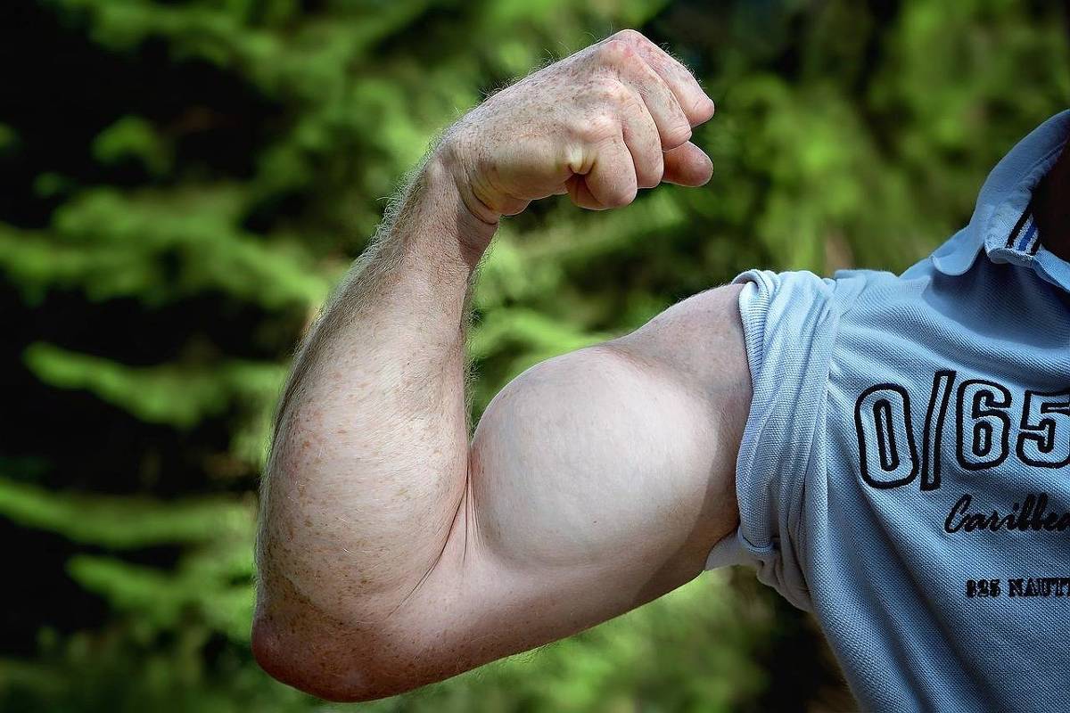 A imagem mostra um braço musculoso de uma pessoa, com o bíceps bem definido, em uma pose de flexão. O braço é de pele clara e está parcialmente coberto por uma camisa de manga curta. Ao fundo, há uma vegetação verde, sugerindo um ambiente ao ar livre.